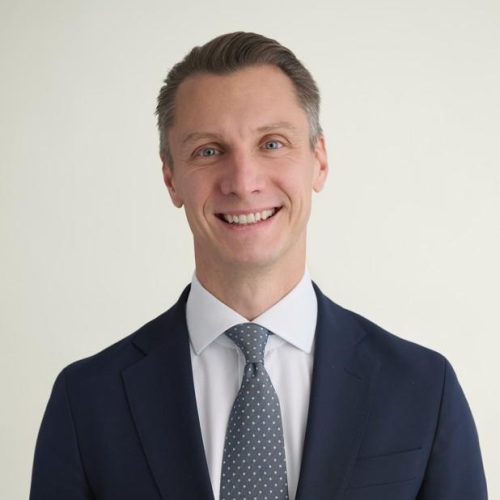 Smiling man in a navy suit and light gray tie, posing for a professional headshot against a pale background.