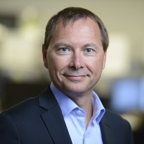 Professional head-and-shoulders portrait of a man in a dark blazer and light blue shirt, smiling at the camera in an office setting.