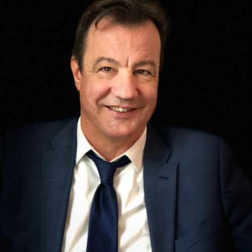 Smiling man wearing a navy suit and blue tie, white shirt, against a dark background.