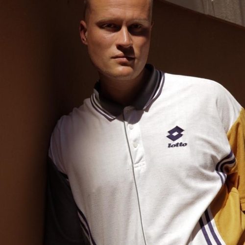 Portrait of a man with short blond hair wearing a white Lotto polo shirt, leaning against a brown wall and looking at the camera.