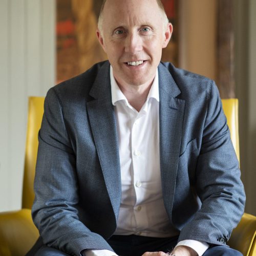 Smiling bald man in a blue suit sits on a yellow chair, hands clasped in his lap indoors.