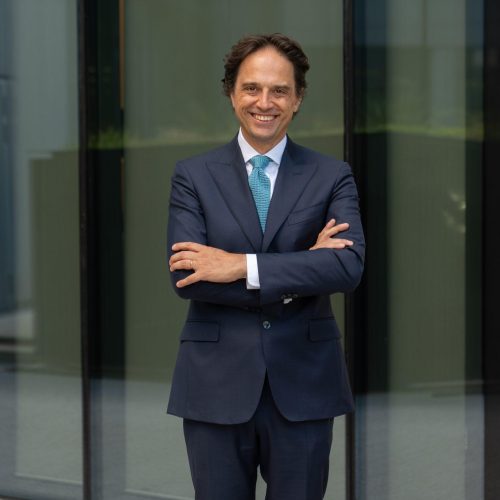 Smiling man in a navy suit and teal tie stands with arms crossed in front of glass building façade.