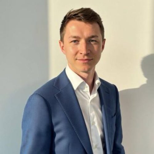 Professional portrait of a man in a blue suit and white shirt, looking at the camera indoors with soft lighting.