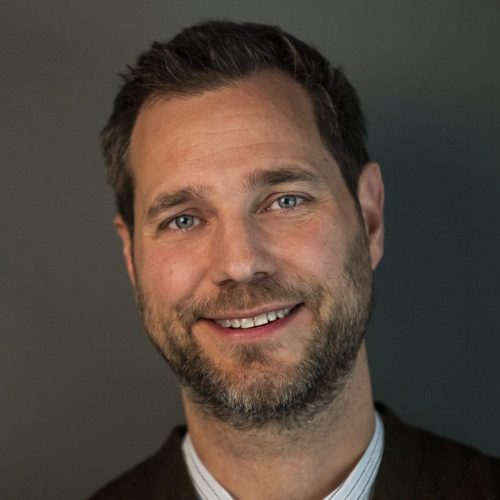 Close-up portrait of a smiling man with blue eyes, short dark hair and light beard, wearing a white shirt and dark blazer.