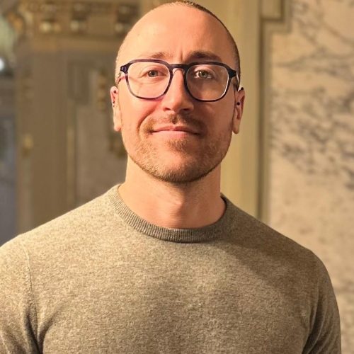 Portrait of a smiling man with short hair and dark-framed glasses, wearing a gray sweater indoors.