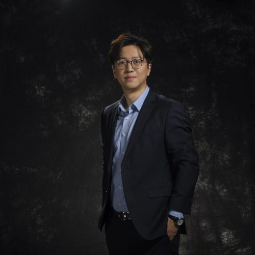Professional man in a dark pinstripe suit and light blue shirt poses against a dark textured backdrop, one hand in pocket, looking at the camera with a slight smile.