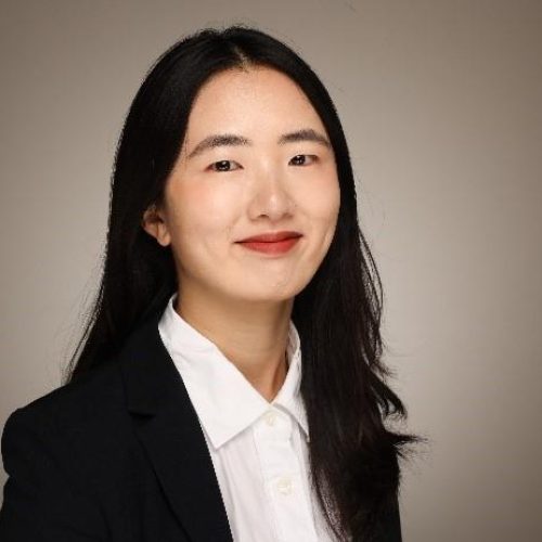 Professional headshot of a woman with long black hair, wearing a black blazer and white shirt, smiling softly against a neutral background.
