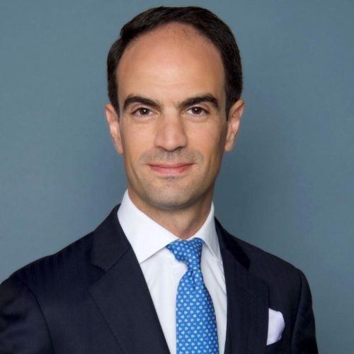 Professional head-and-shoulders portrait of a man in a navy suit and blue tie in front of a blue background.